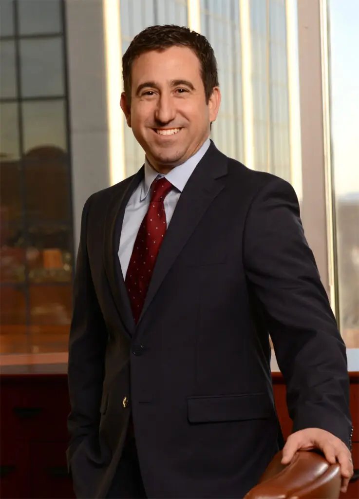 Man in a dark suit, light blue shirt, and red patterned tie smiling