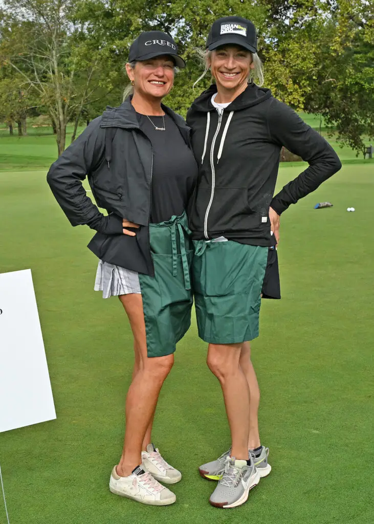 2 woman standing on a golf course wearing black ball caps and green aprons