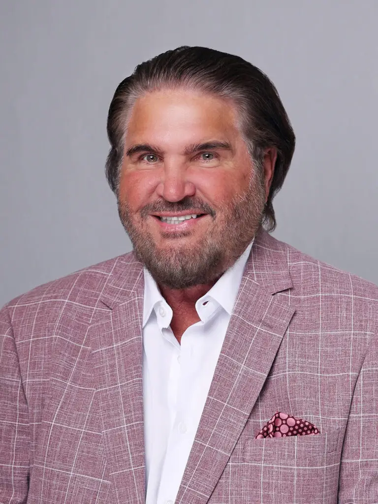A dark haired man with a beard, wearing a pink sports jacket with a white window pane pattern and a pocket handkerchief