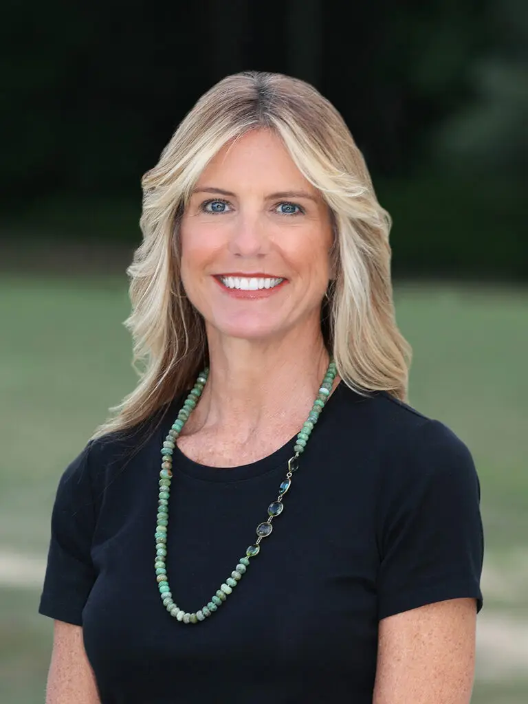 A blond haired woman, against an outdoor background ,wearing a black shirt with a turquoise necklace smiling.